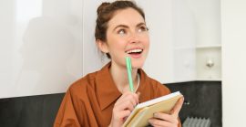 Portrait Of Young Smiling Woman With Creative Thoughts, Looking Up With Inspired Face, Holding Pen And Notebook, Writing Down Her Ideas